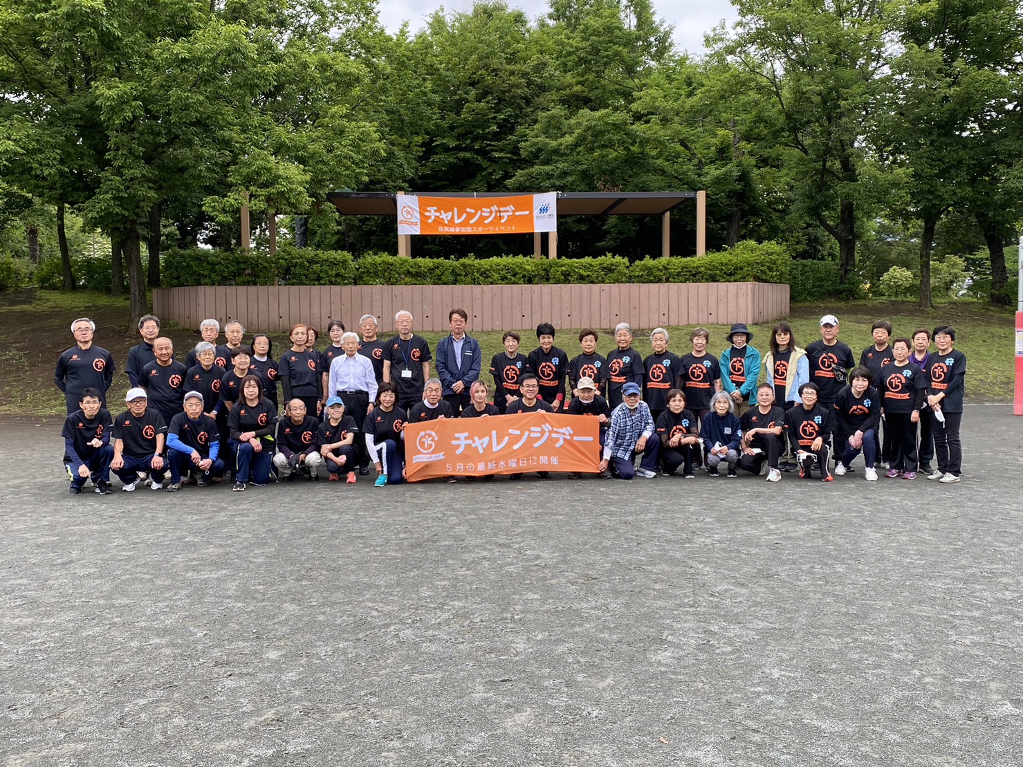 青葉が茂る公園で、お揃いの黒いTシャツを着た数十人の老若男女が、オレンジ色の「チャレンジデー」と書かれた横断幕を掲げて整列している集合写真