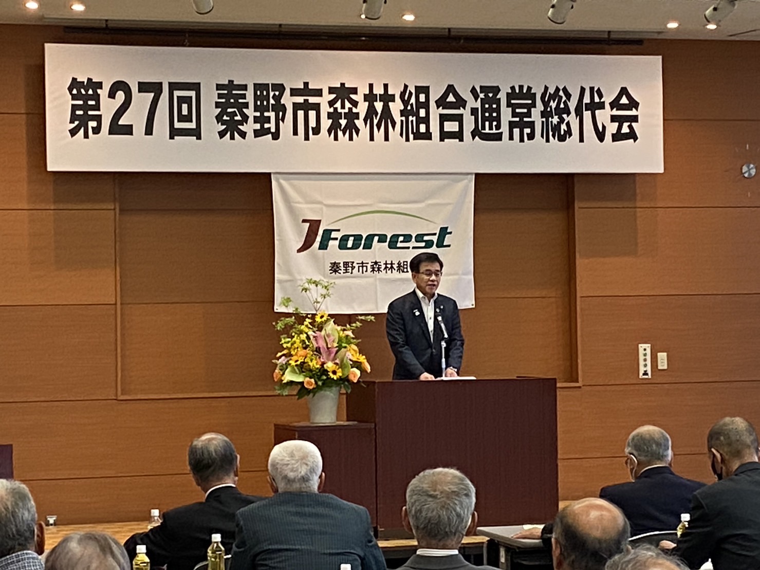 茶色い壁の壇上で、男性がマイクに向かってスピーチをしており、その上には「第27回秦野市森林組合通常総代会」の垂れ幕がかかっている様子の写真