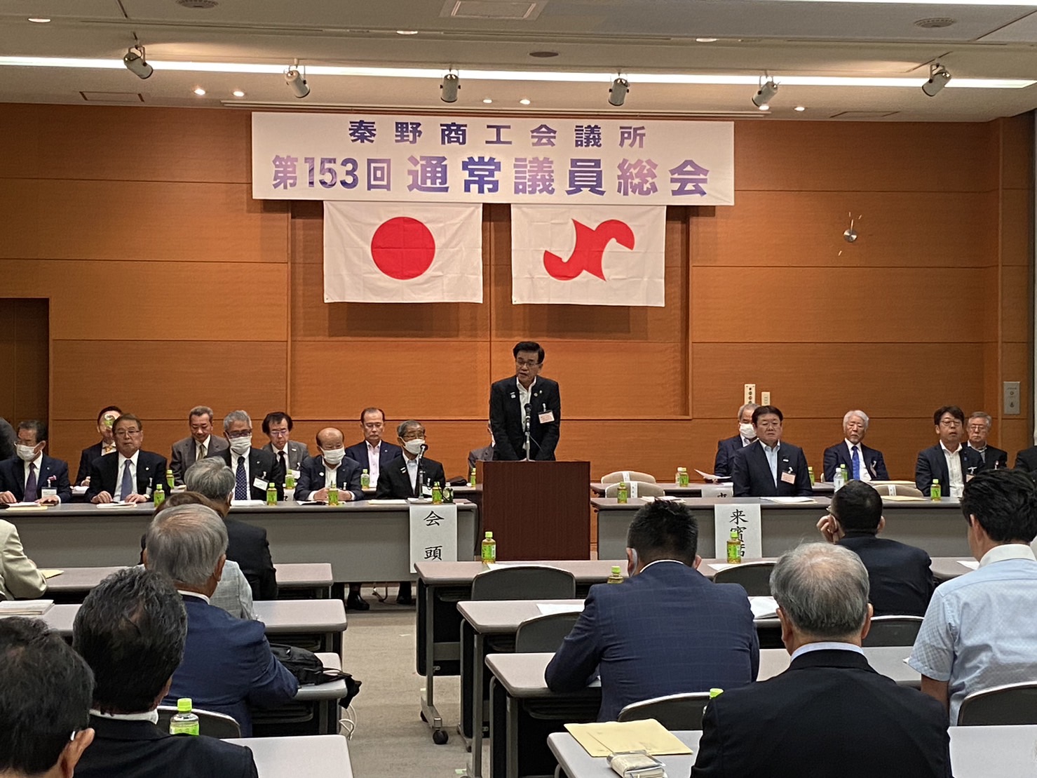壇上に「第153回通常議員総会」の垂れ幕と日本国旗などが飾られている和やかな雰囲気の室内で、演台に立つ市長がスピーチをしている写真