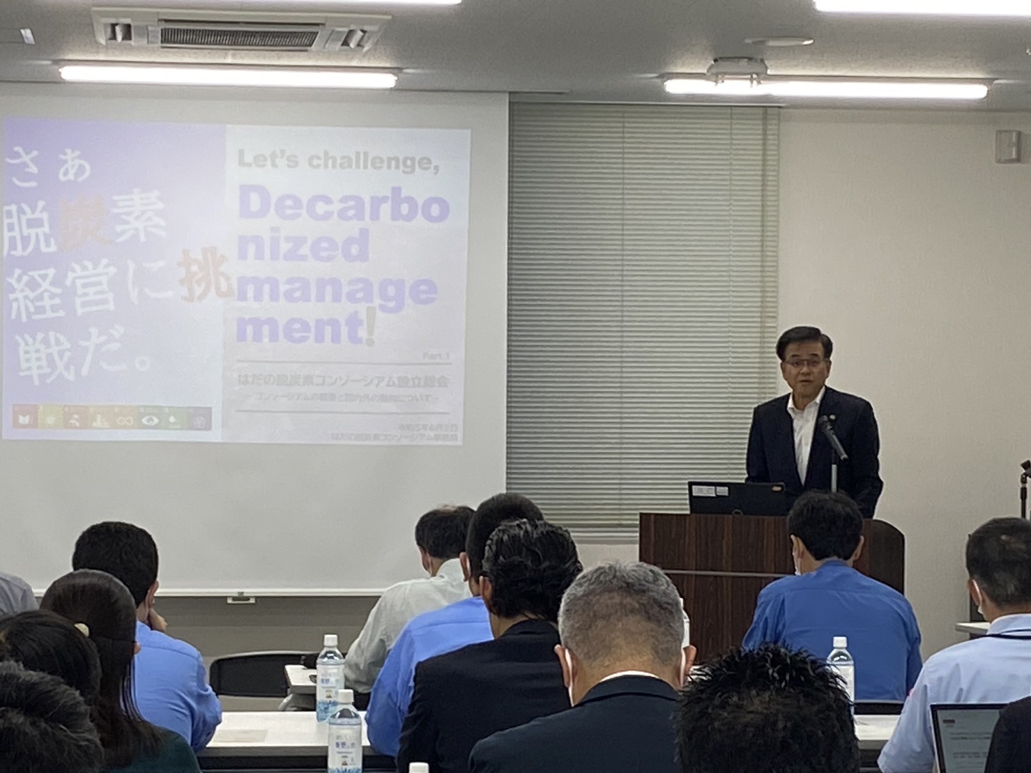 演台に立つ市長が、スクリーンを背景に、多くの人々を前にスピーチをしている様子の写真