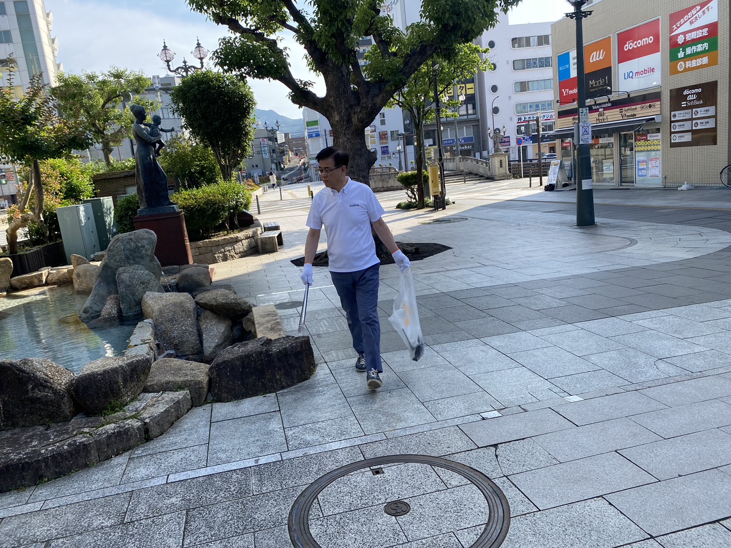 白いポロシャツに軍手をはめた市長が、トングとゴミ袋を持ち、舗道に設置された噴水のそばで清掃活動をしている様子の写真