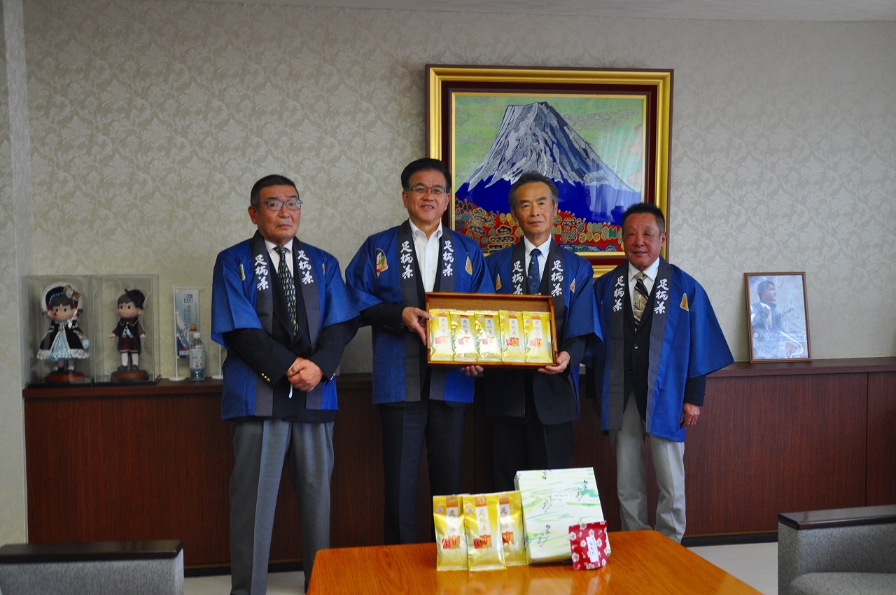 「足柄茶」と書かれた青い法被を着用した4人の男性が、富士山の絵画を背景に、お茶がはいった箱を持ち、机にお茶や箱などの品物が並べられた集合写真