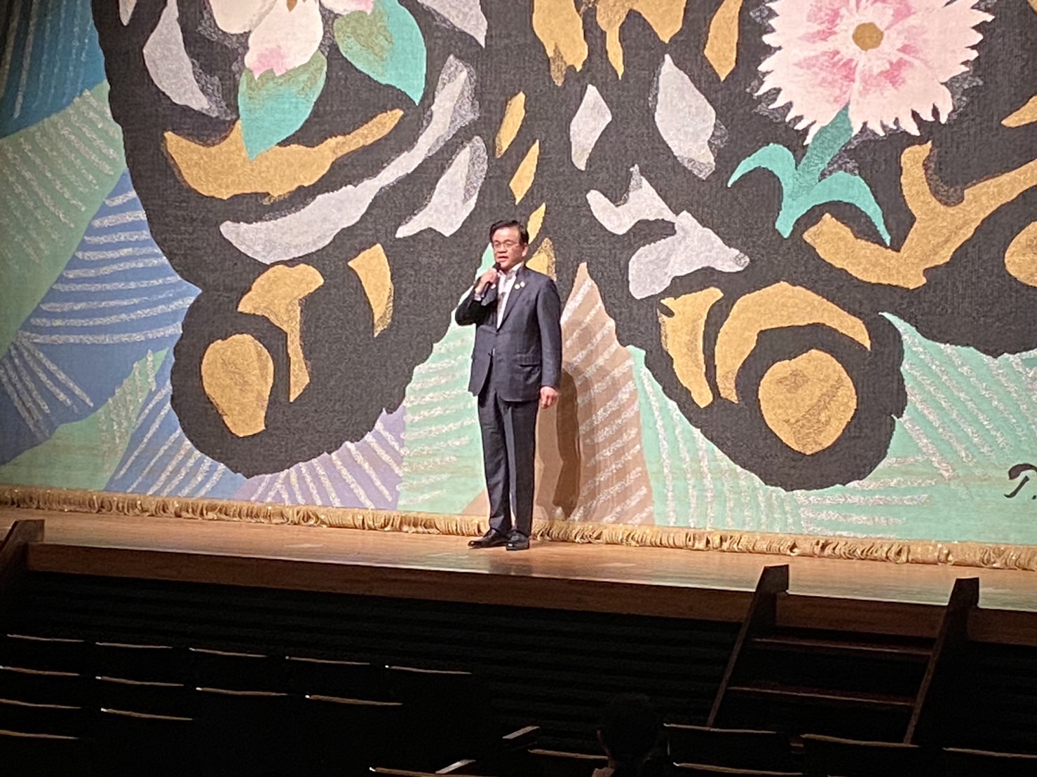 華やかな色彩のタペストリーを背景にしたステージで、スーツ姿の市長がマイクを持って聴衆にスピーチをしている様子の写真