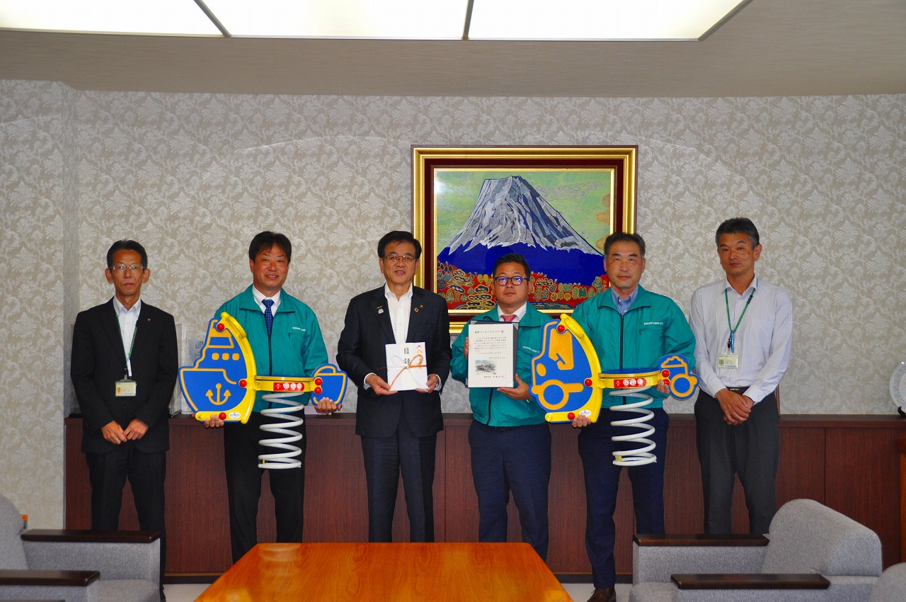 和やかな雰囲気の室内で、富士山の絵画を背景にスーツ姿の男性3人と作業着姿の男性3人が、記念品を持って並んでいる様子の集合写真