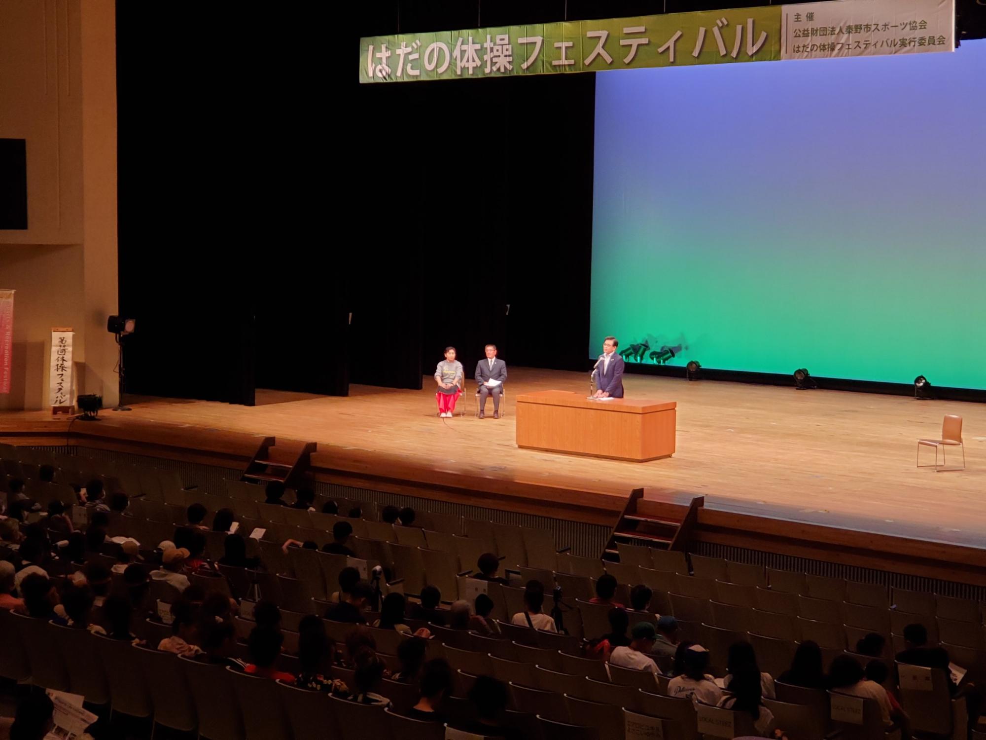 大きな体育館のステージ上で、真ん中に座る2人の人物と、演台で挨拶をする市長がおり、客席には多くの観客が着席している様子の写真