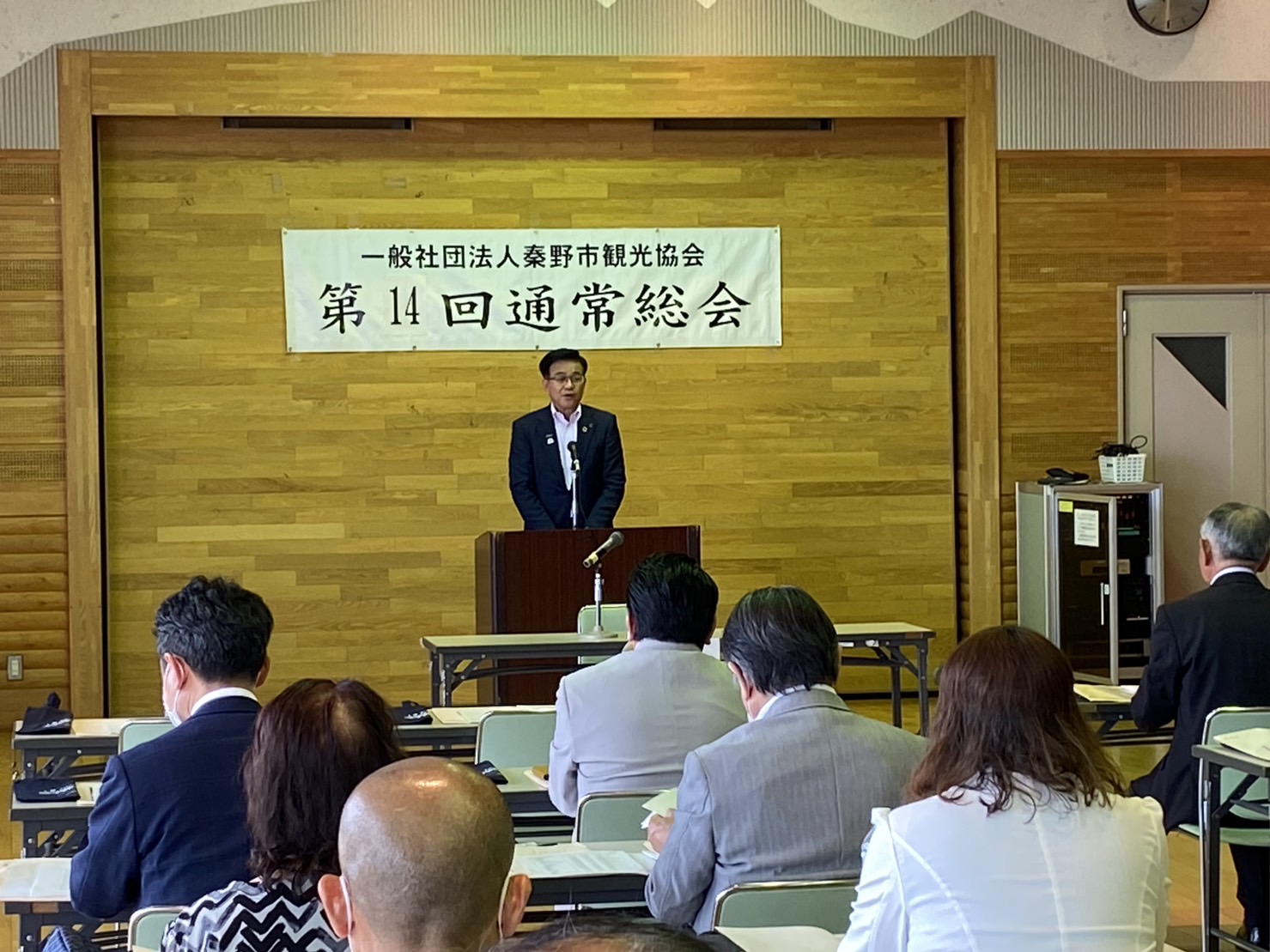 木目調の壁を背景に、「一般社団法人秦野市観光協会 第14回通常総会」と書かれた横断幕の下で、市長が演台から挨拶をしており、それを多くの人々が聞いている様子の写真