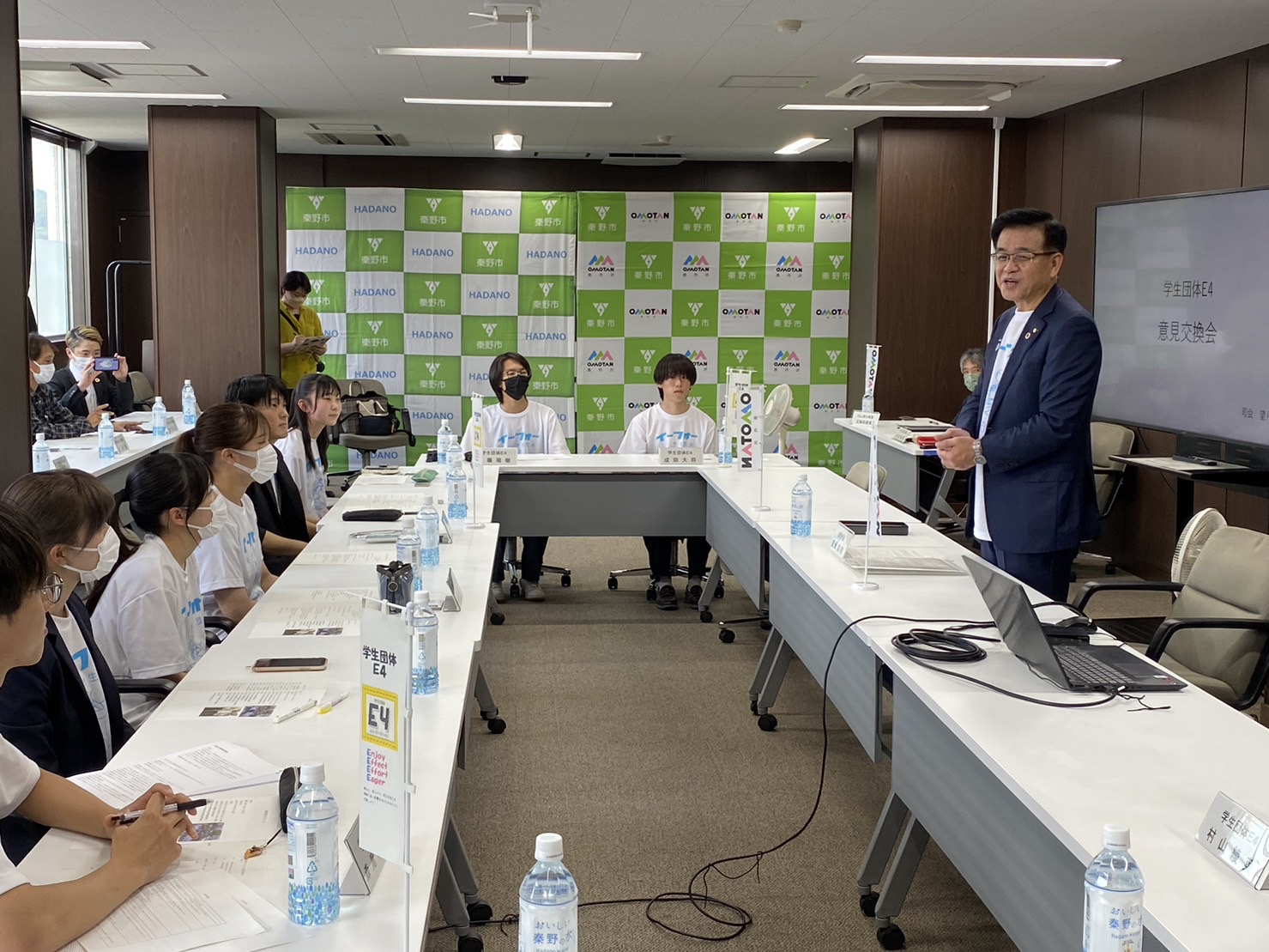 「HADANO秦野市」と書かれたバックパネルが飾られた室内で、白い長机に座った学生たちに向けてスーツ姿の市長が話をしている写真