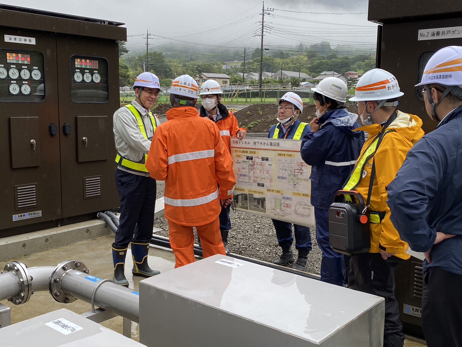 ヘルメットをかぶり、白にオレンジのラインの入った作業服を着た複数の作業員が、機械が設置された屋外で、図面を囲んで真剣に話し合っている様子の写