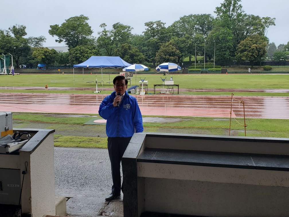 青いジャンバーを着た市長が雨のグランドをバックにマイクを使い挨拶をしている写真