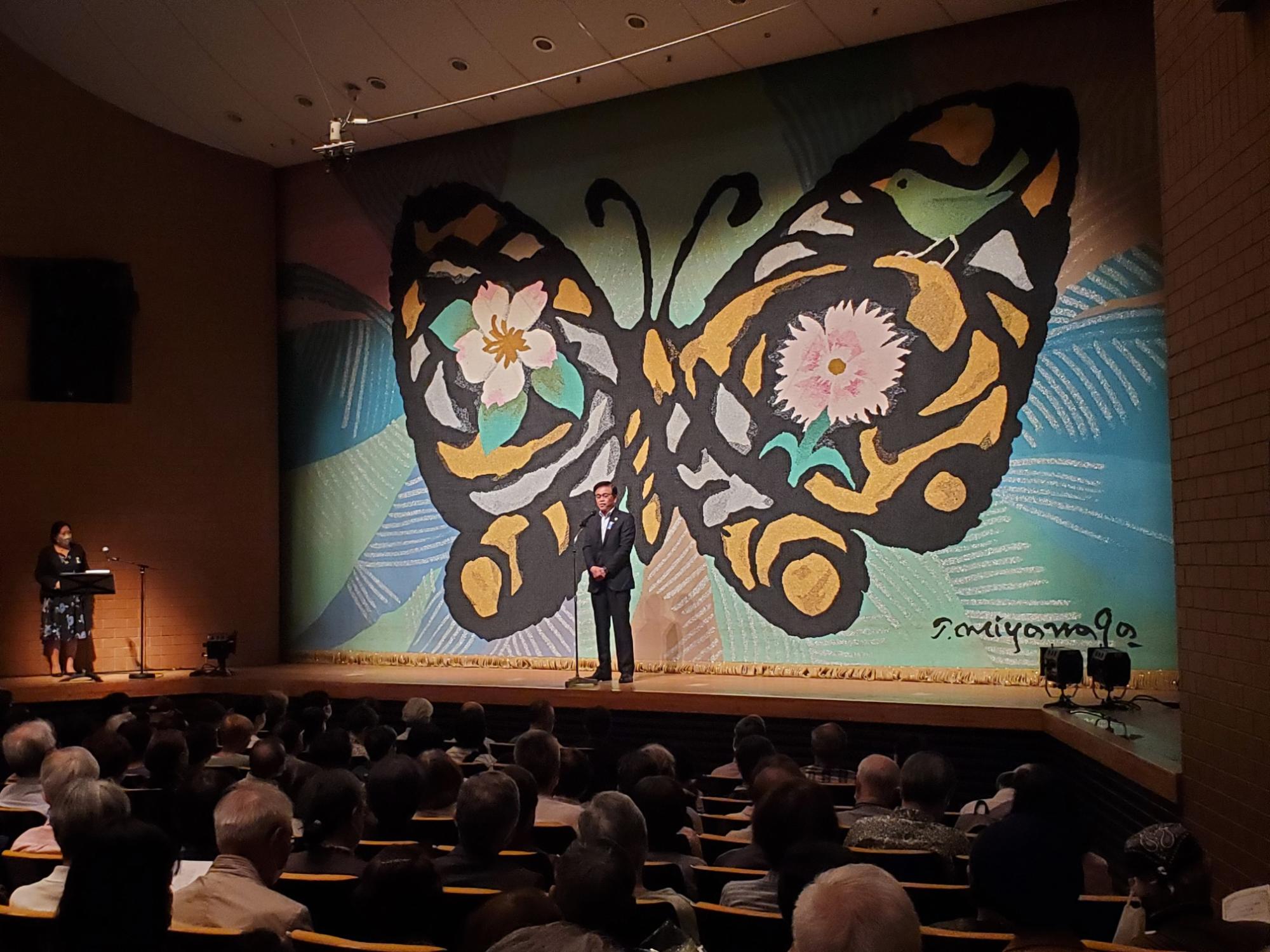 蝶のイラストが描かれた舞台の幕の前でスーツ姿の市長が挨拶をしている様子を写した写真