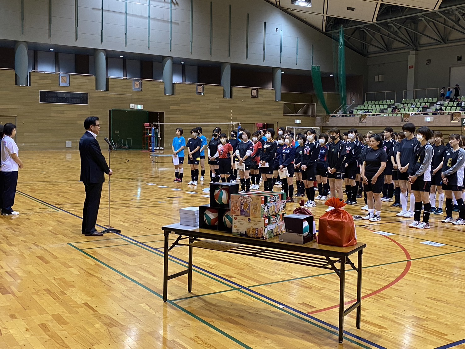 手前の机には記念品が並べられている体育館に立ったスーツ姿の市長が、多くのスポーツ選手たちに向かってスピーチをしている写真