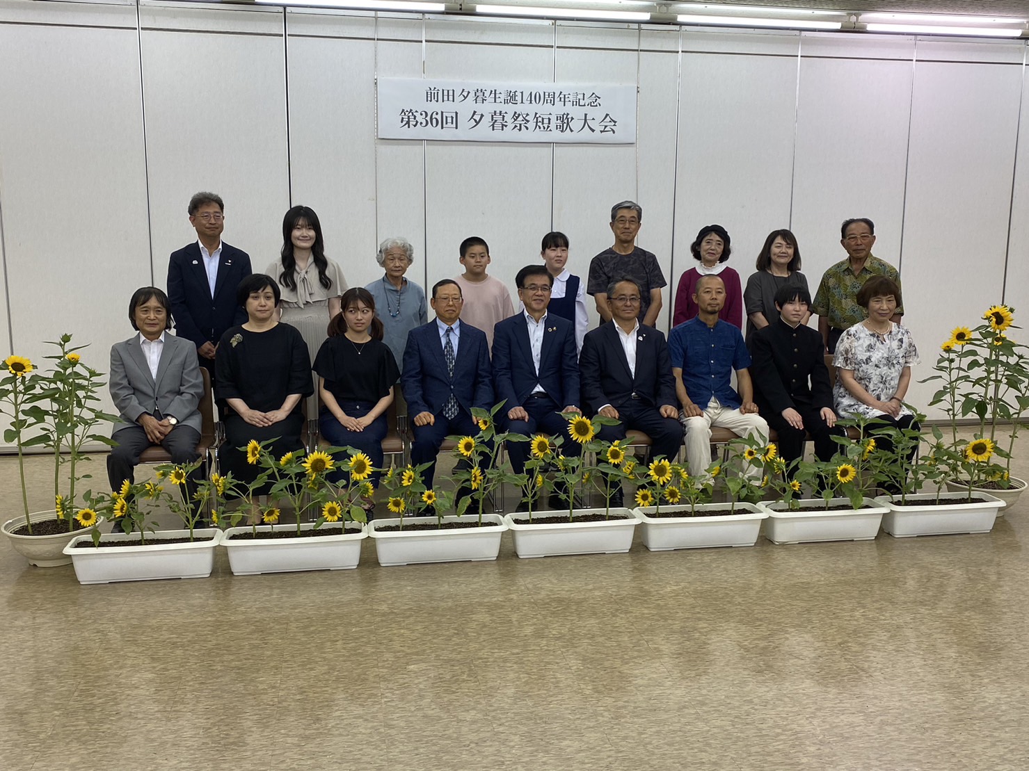 「前田夕暮生誕140周年記念第36回夕暮祭短歌大会」と書かれた横断幕の下で、前列中央に座ったスーツ姿の市長を中心に左右と後方に分かれて老若男女が並び、前にはプランターに植えられたヒマワリが置かれている集合写真