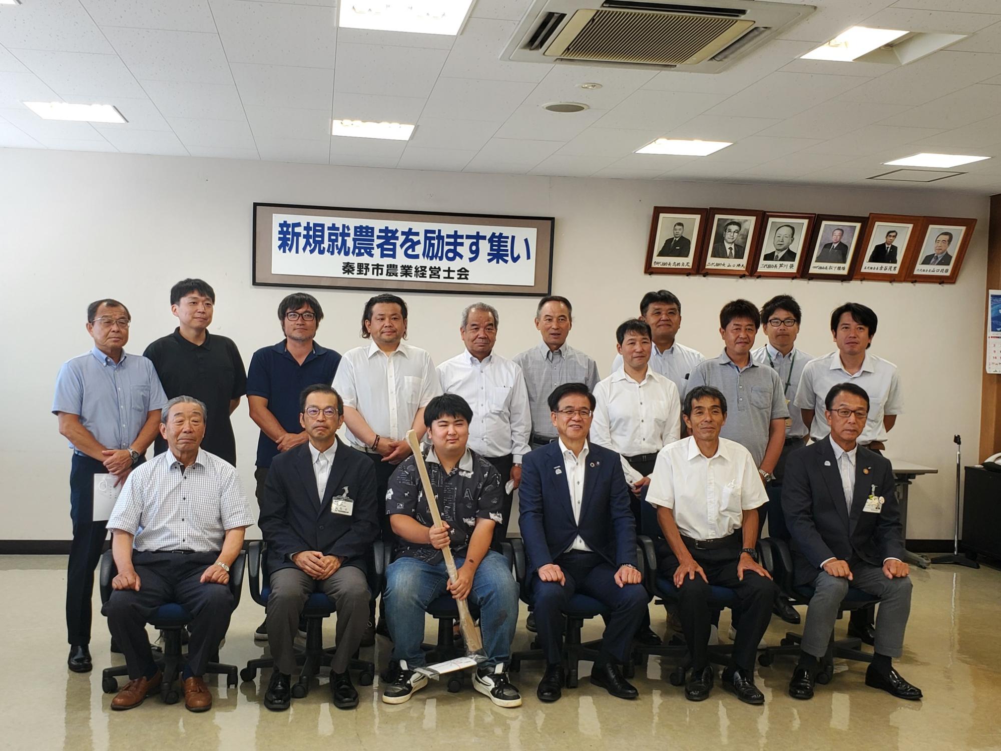 横長の額に入った「新規就農者を励ます集い」と書かれた横断幕の下で、スーツ姿の市長と平鍬を持った若い男性を中央に、左右と後方に分かれて老若男女が並び、全員がカメラ目線で笑顔を向けている集合写真