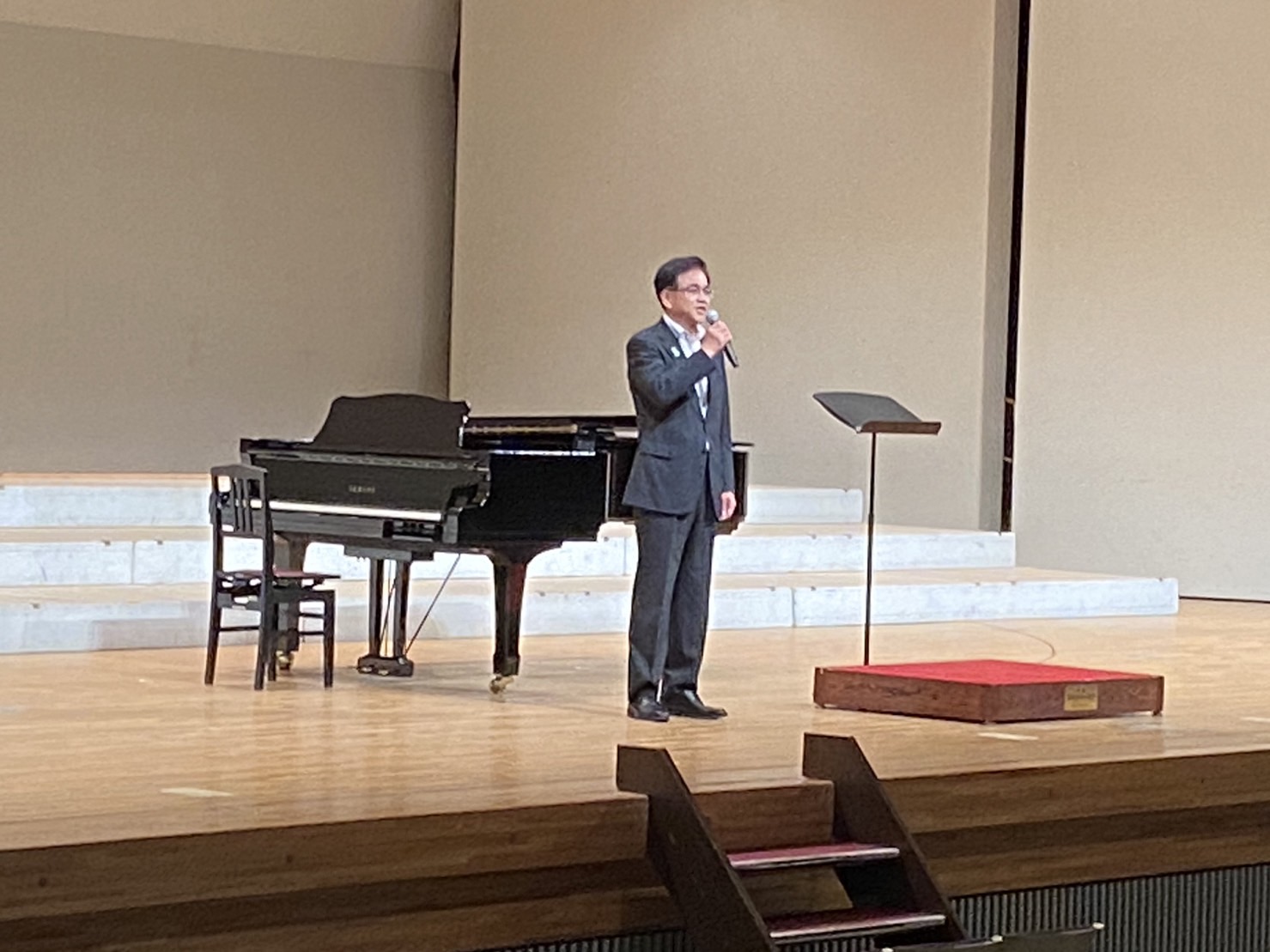 中央にグランドピアノ、指揮台と譜面台が置かれた白い背景のステージの上で、右手にマイクを持ち立っているスーツ姿の市長が挨拶をしている様子を撮影した写真