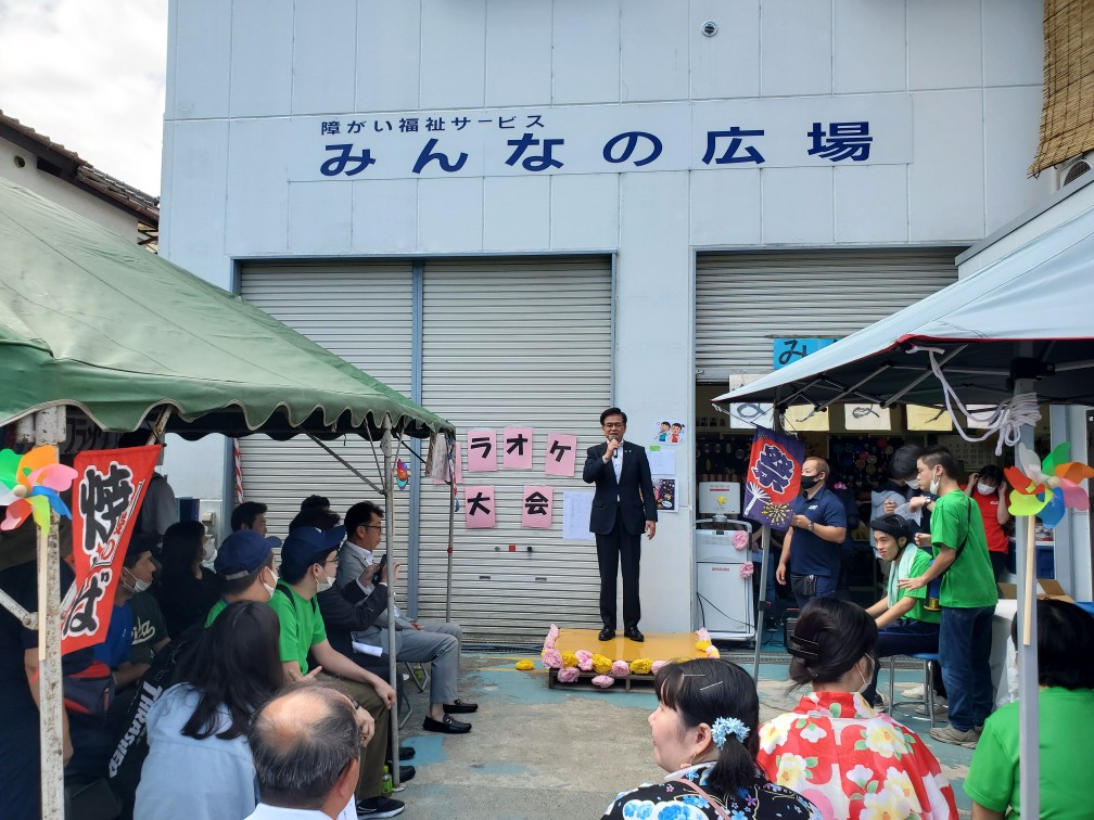 「障がい福祉サービスみんなの広場」と書かれた白壁の建物の前で、右手にマイクを持ったスーツ姿の市長が小型の舞台に立ち挨拶をしており、その周りでは緑色のシャツを着たスタッフや浴衣姿の女性など多くの人々がテントの下に座って話を聞いている様子の写真