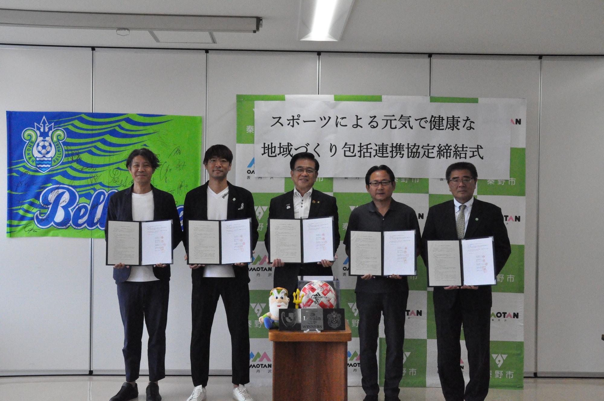 白壁に旗が飾られ、「スポーツによる元気で健康な地域づくり包括連携協定締結式」と書かれた秦野市のバックパネルの前で、スーツ姿の市長を中央に4人の男性が、それぞれ協定書を持って並び、全員がカメラ目線で立っている記念写真