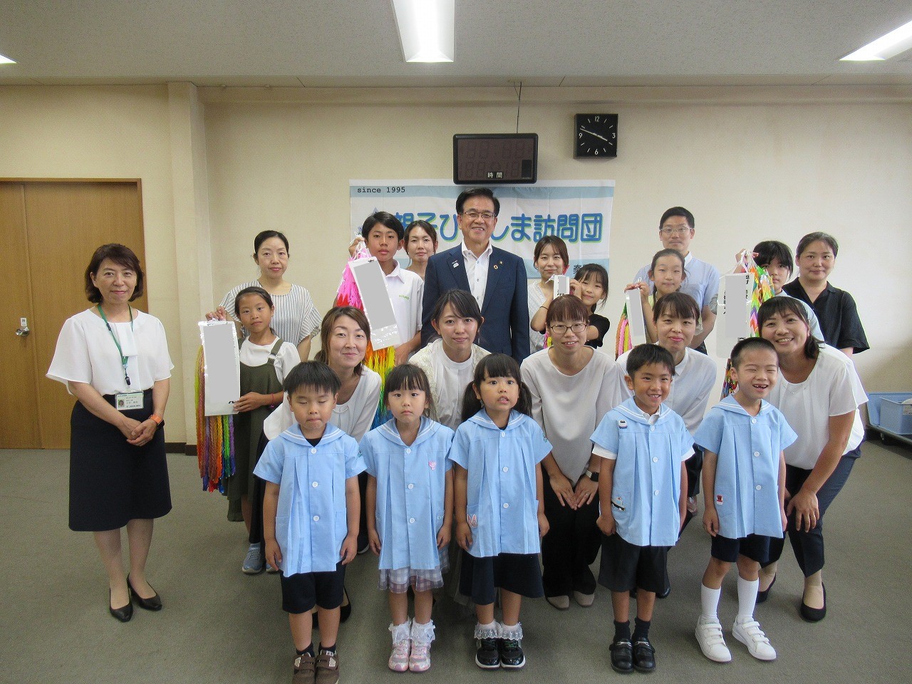公共施設の部屋で、「親子ひまわり訪問団」と書かれた白壁を背景に、水色の制服を着用している子供や大人まで、様々な年齢層の男女が全員カメラ目線で並んでいる集合写真