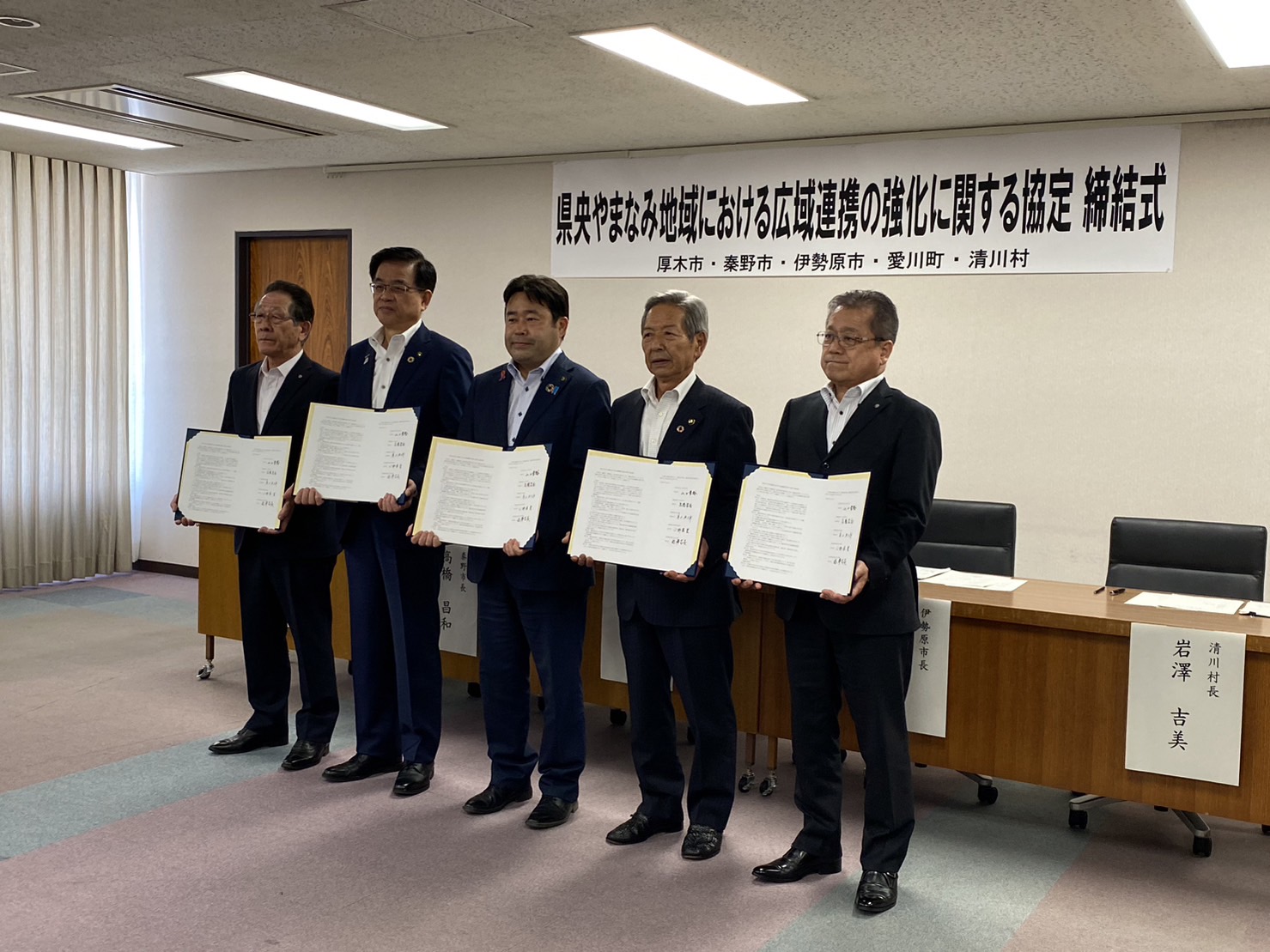 「県央やまなみ地域における広域連携の強化に関する協定 締結式」と書かれた横断幕が貼られた室内で、5人の男性がそれぞれの協定書を持ってカメラに向かって並んでいる記念写真