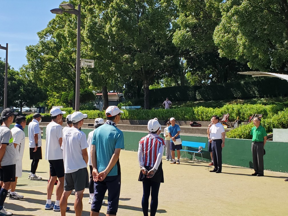 周囲を大きな木々や生垣に囲まれた広々としたテニスコートで、白い帽子とウェアを身につけた複数の選手が整列しており、その向かいで白いシャツを着た市長がマイクを持って挨拶をしている様子を捉えた写真