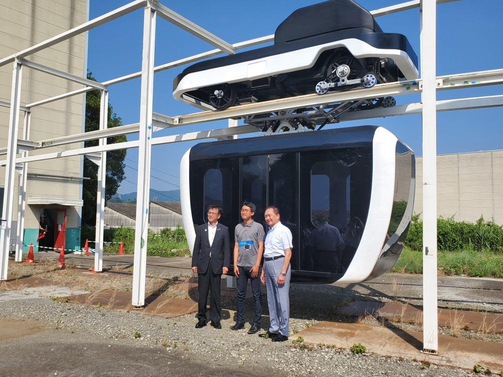 白壁の建物前の屋外で、黒と白を基調とした未来的なデザインのロープウェイの様な乗り物の前で、スーツ姿の男性を含む3人が立っている様子を撮影した写真