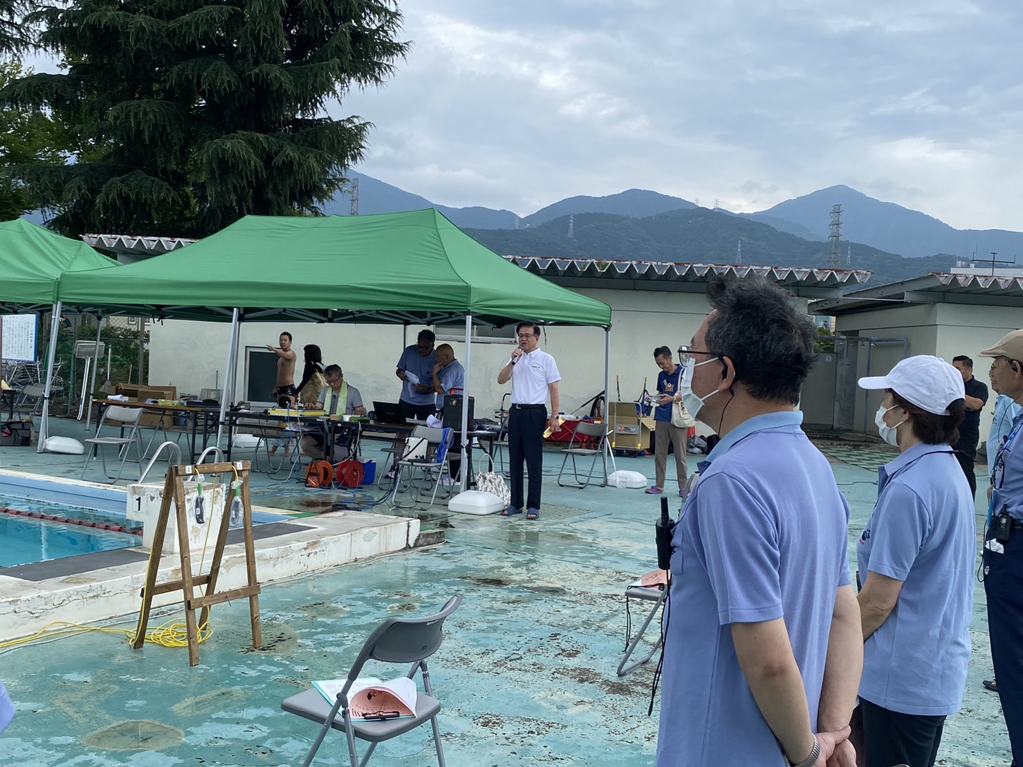 山の景色が見える屋外プールのプールサイドに立つお揃いの青色のシャツを着たスタッフや、緑色のテントの下にいるスタッフの方々が、テントの傍に立っている白いシャツを着た市長がマイクを持って話している様子を聞いている写真