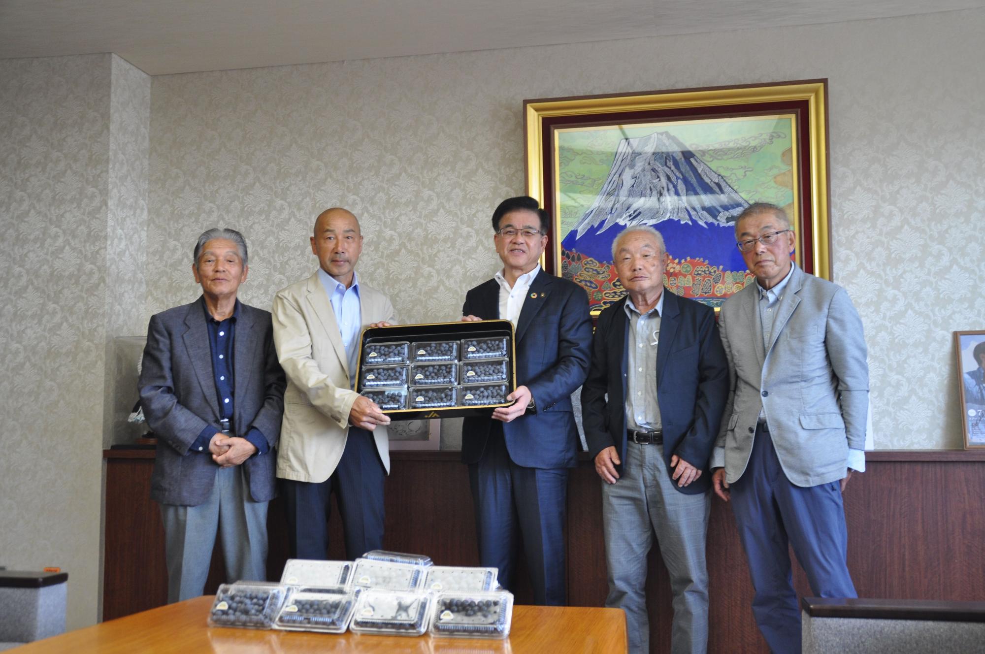 金色の額縁に入った山頂に雪が積もった大きな山の絵画が壁に飾られた落ち着いた背景の部屋で、5人の男性が並んでおり、左から2番目に立つ男性と中央の市長がブルーベリーが入ったパックが並んだ盆を両手で一緒に持っている集合写真