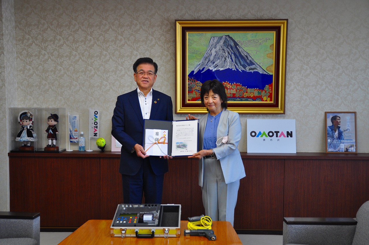 金色の額縁に入った山頂に雪が積もった大きな山の絵画が壁に飾られた落ち着いた背景の市役所の部屋で、スーツ姿の市長と女性が並んでおり、2人で目録の封筒が入った協定書のような書類をカメラに向けて持っている記念写真