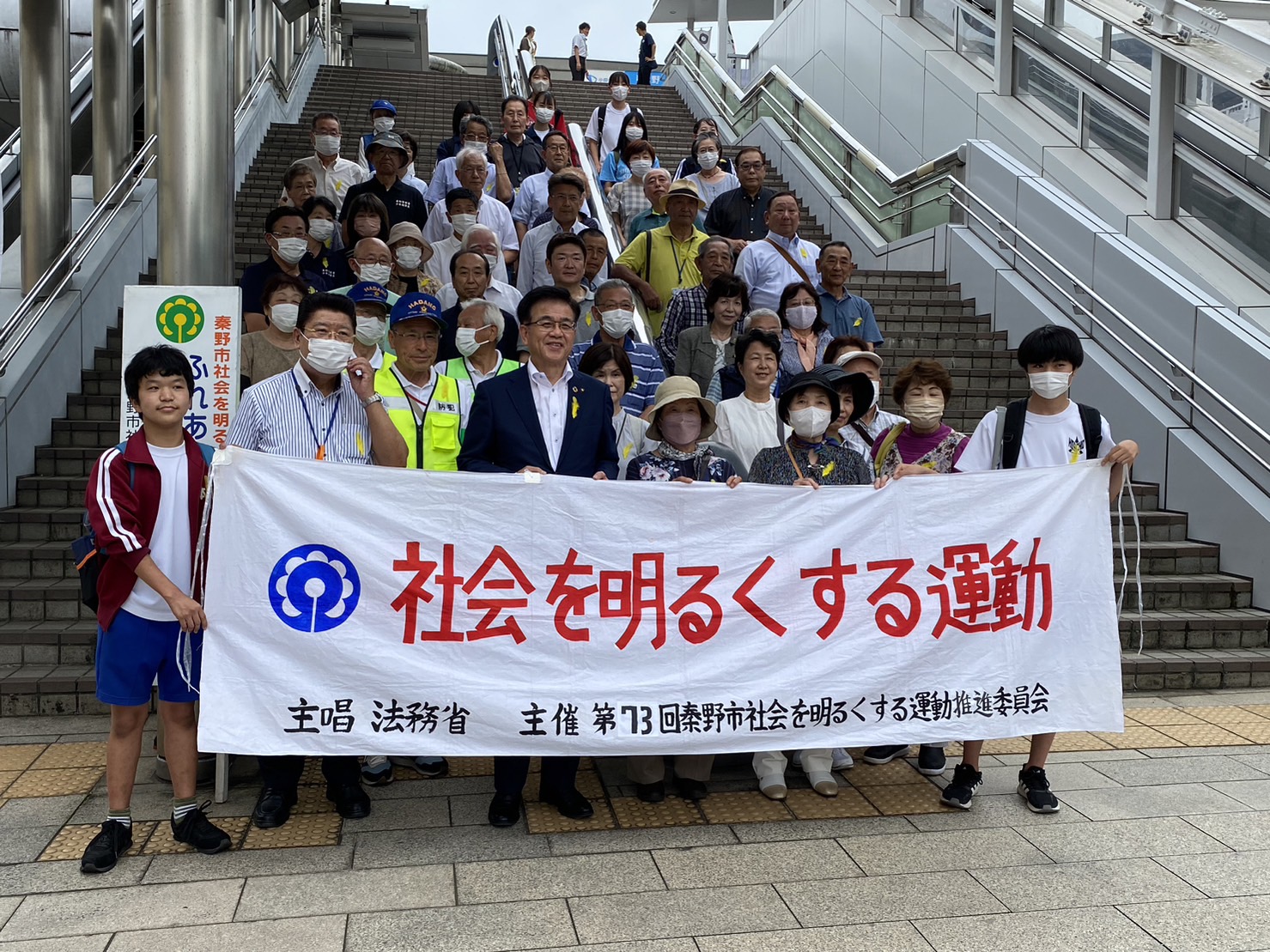 「社会を明るくする運動」と書かれた横断幕を先頭に、スーツ姿の市長やマスクを着用した多くの人々が駅の広い階段に並んでいる様子の写真