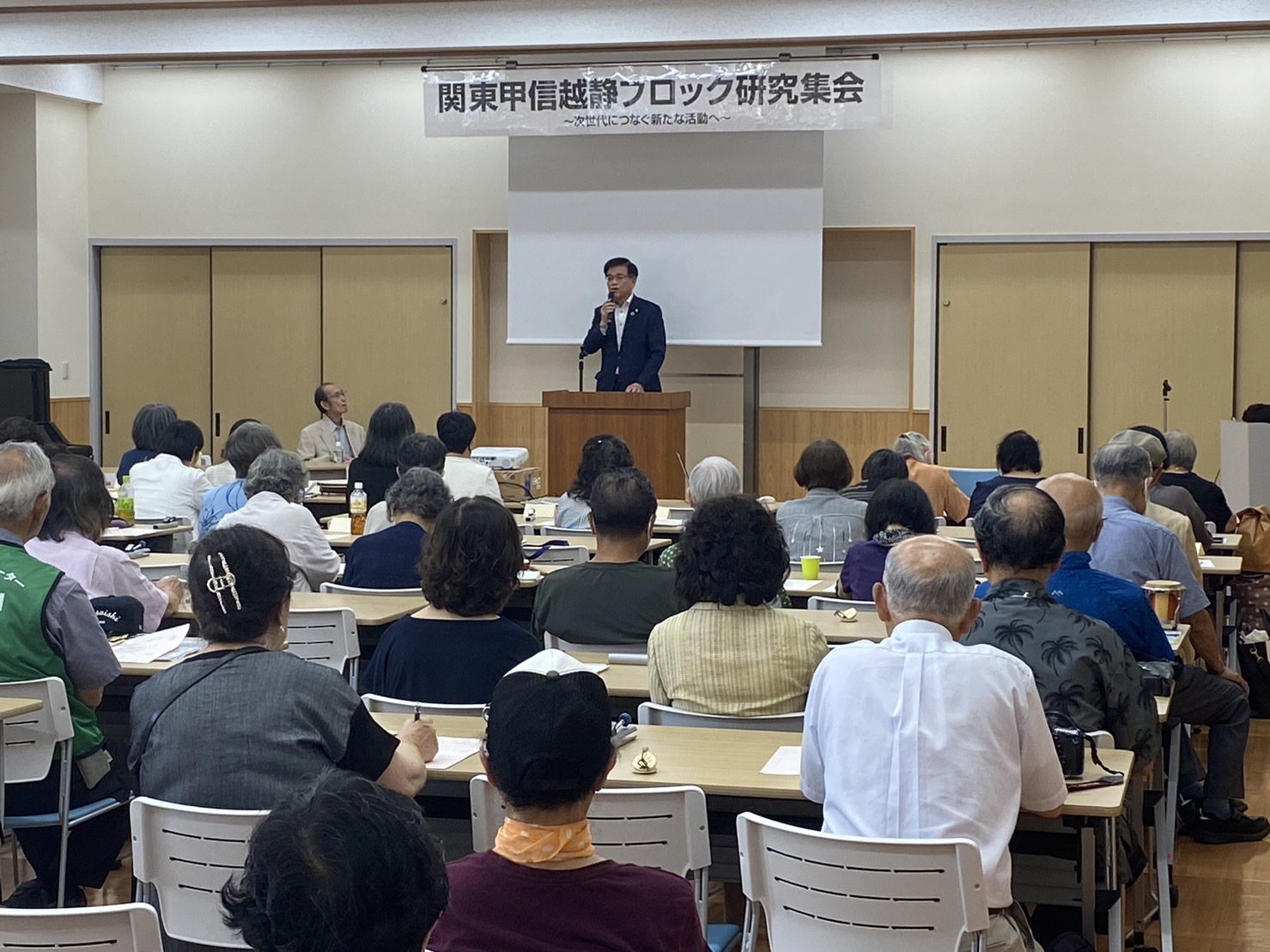 「関東甲信越静ブロック研究集会」と書かれた横断幕とスクリーンの前で、スーツ姿の市長が演台に立って話をしており、多くの参加者が熱心に耳を傾けている様子の写真