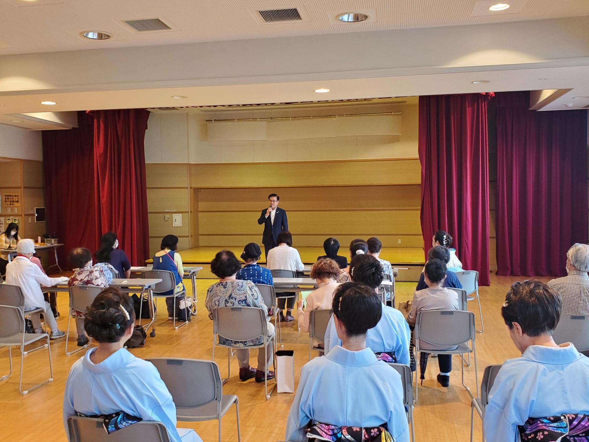 小さなステージのある室内で、赤色のカーテンを背景にスーツ姿の市長がマイクを持って挨拶をしており、その話を聞くために集まった多くの人々が椅子に座っている様子の写真