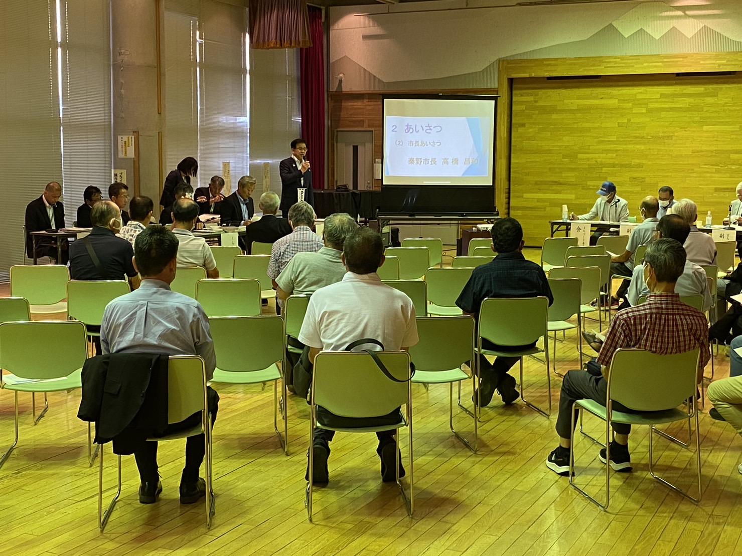 公共施設の広々としたホールの前方に設置されたスクリーンの左側に、スーツ姿の市長が挨拶をしており、その話を聞くために集まった多くの人々が等間隔に並べられた椅子に座っている本町地区で行われた市政懇談会の様子の写真