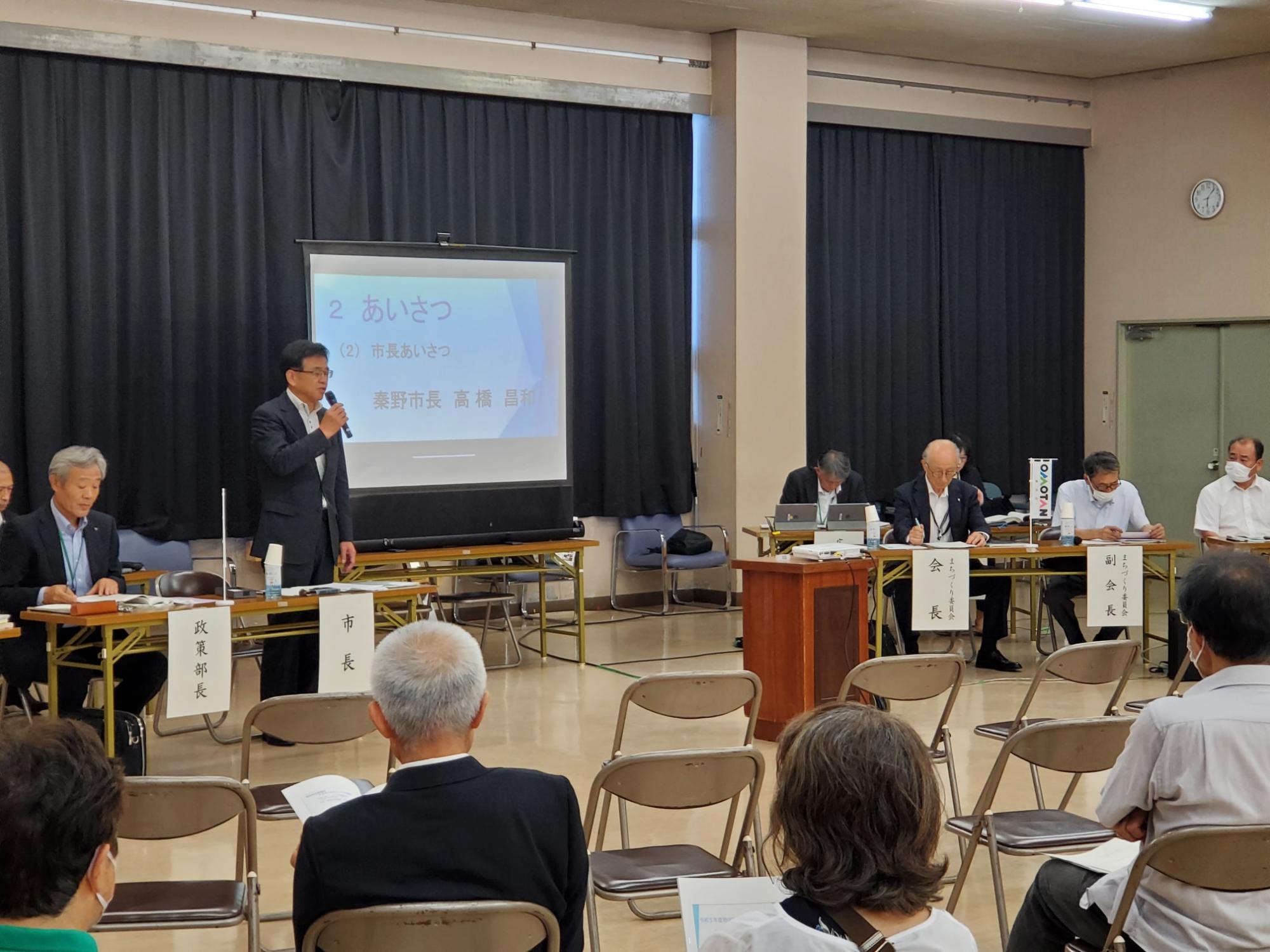 黒いカーテンがひかれた公共施設のホールに「あいさつ」と表示されたスクリーンが設置され、左側の席から立ち上がったマイクを持って話す市長の話を、関係者やパイプ椅子に座った参加した人々が聞いている大根地区で行われた市政懇談会の様子の写真