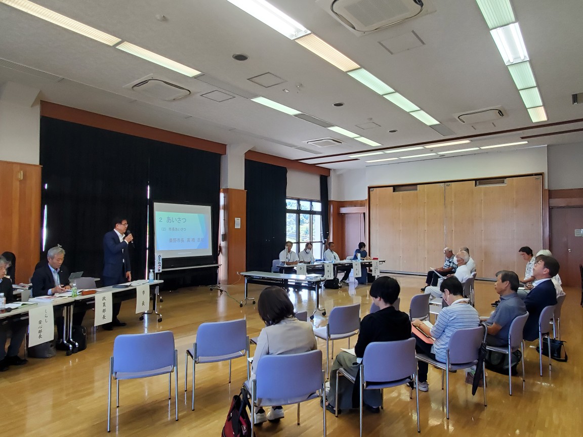 公共施設の広々としたホールの前方に設置されたスクリーンの左側の席から立ち上がったスーツ姿の市長が挨拶をしており、その周りには多くの関係者や参加者が椅子に座って話を聞いている上地区で行われた市政懇談会の様子の写真