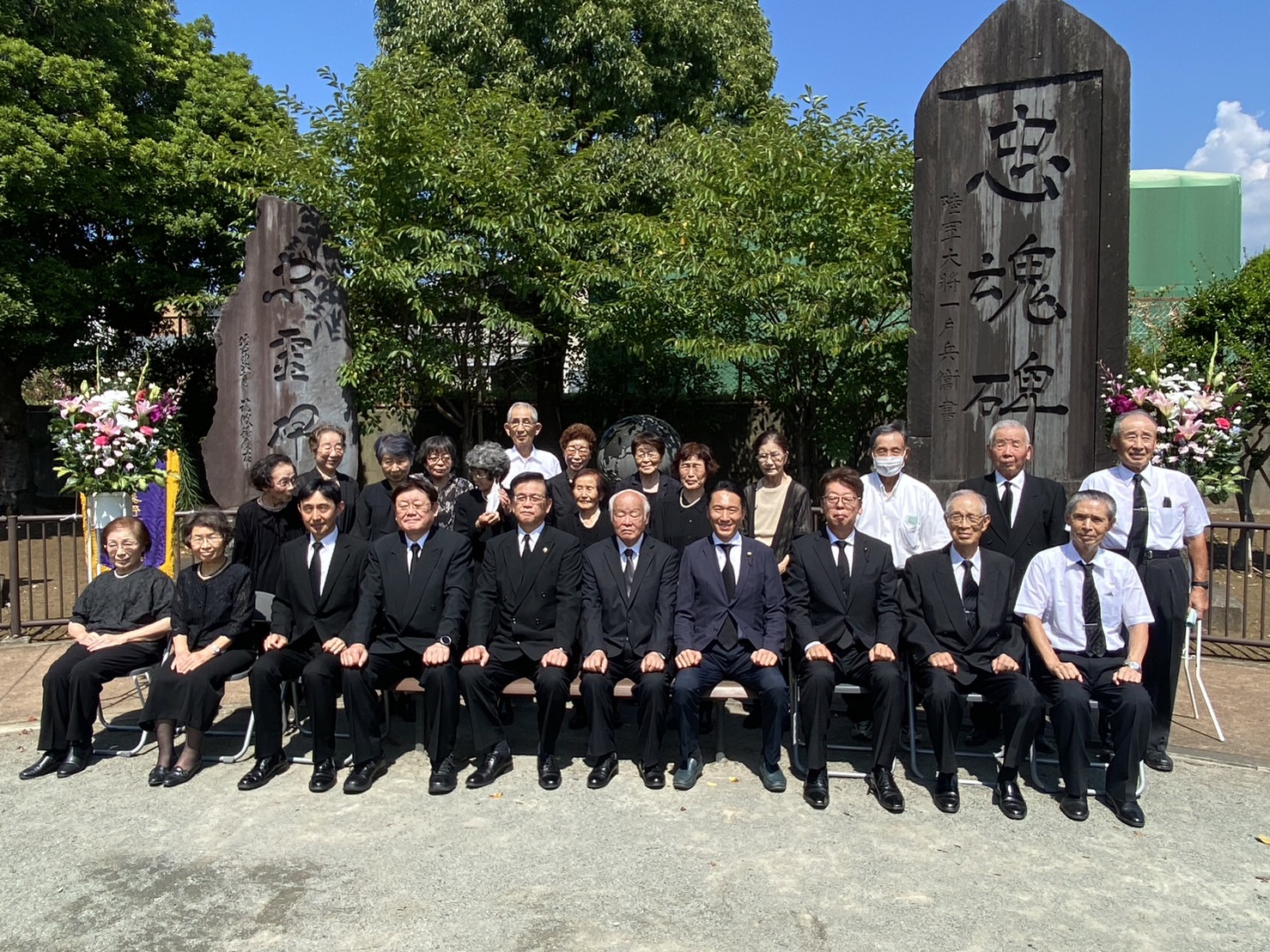 たくさんの人々が黒い服やスーツを着て、墓石の前で並んで座り、その後ろには立ち姿の男女もいる集合写真