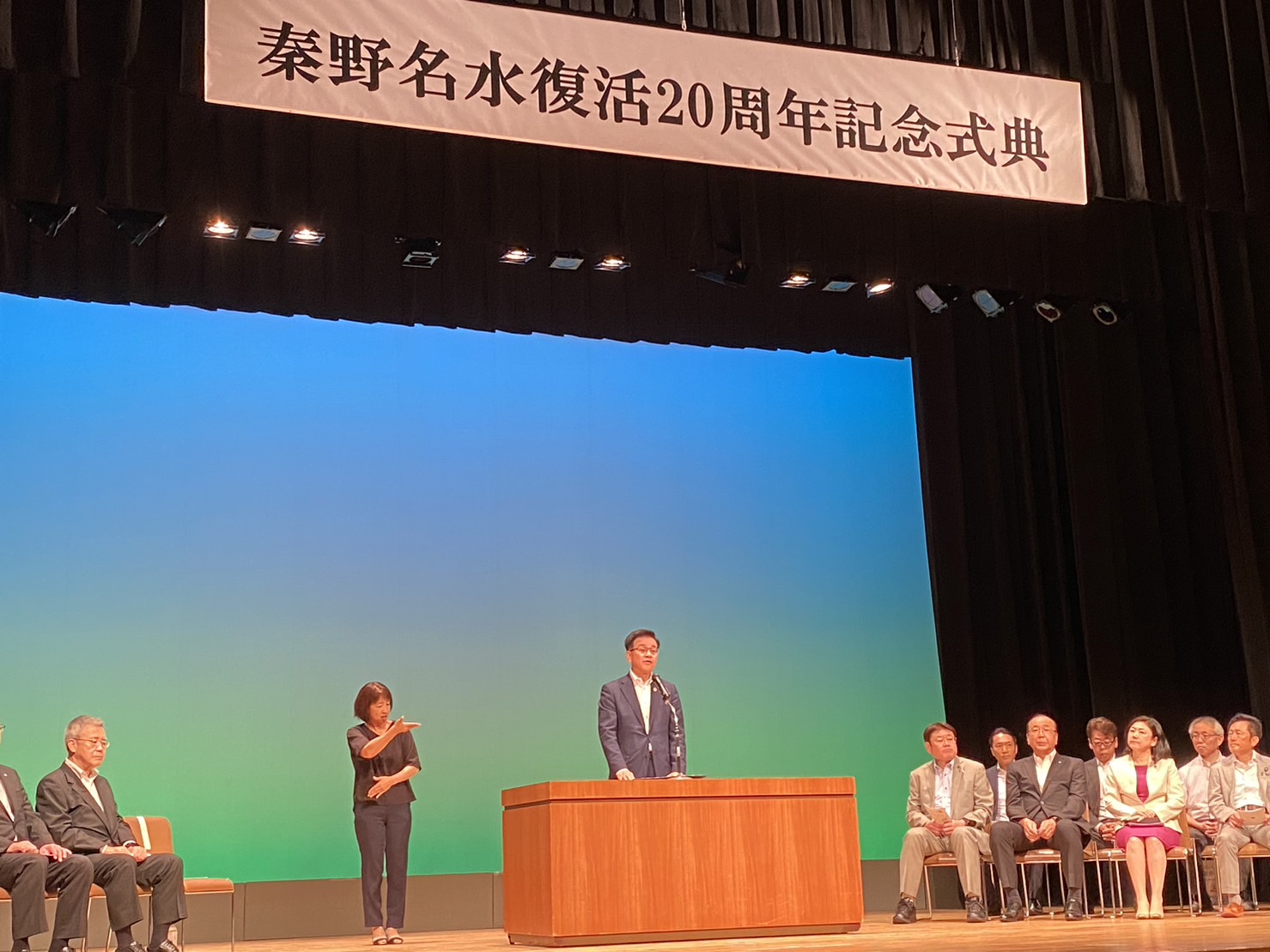 「秦野名水復活20周年記念式典」の横断幕が飾られ、青いスクリーンを背景にしたステージで、演台に立つ市長がスピーチをしており、周りには聴衆と手話通訳を行う女性が立っている様子の写真