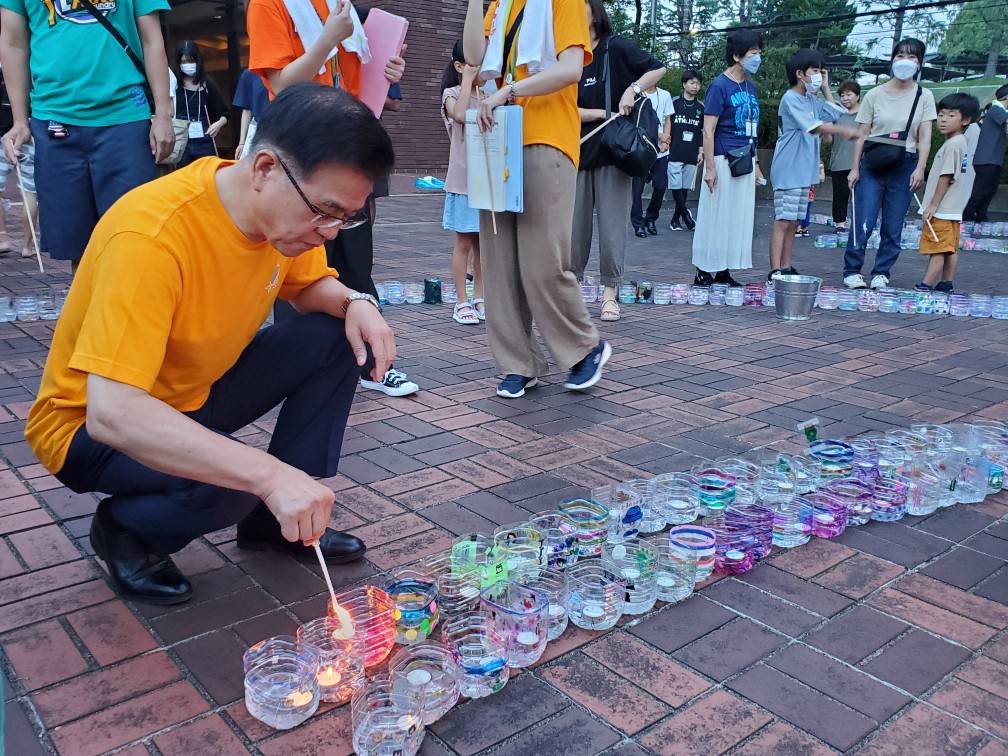 周りには多くの見物人がいる中、広場の舗道でオレンジ色のTシャツを着た市長がしゃがみ込み、並べられたプラスチック製の容器に入ったロウソクに火を灯している写真