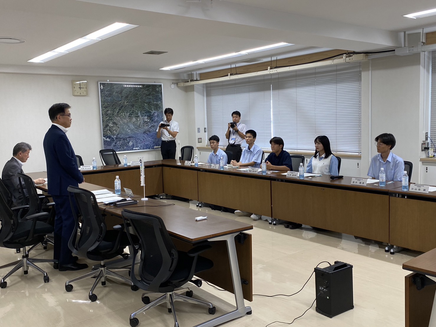 複数の人がコの字型に配置された会議机に座っており、スーツ姿の市長が中央に立って学生たちと向かい合っている様子の写真
