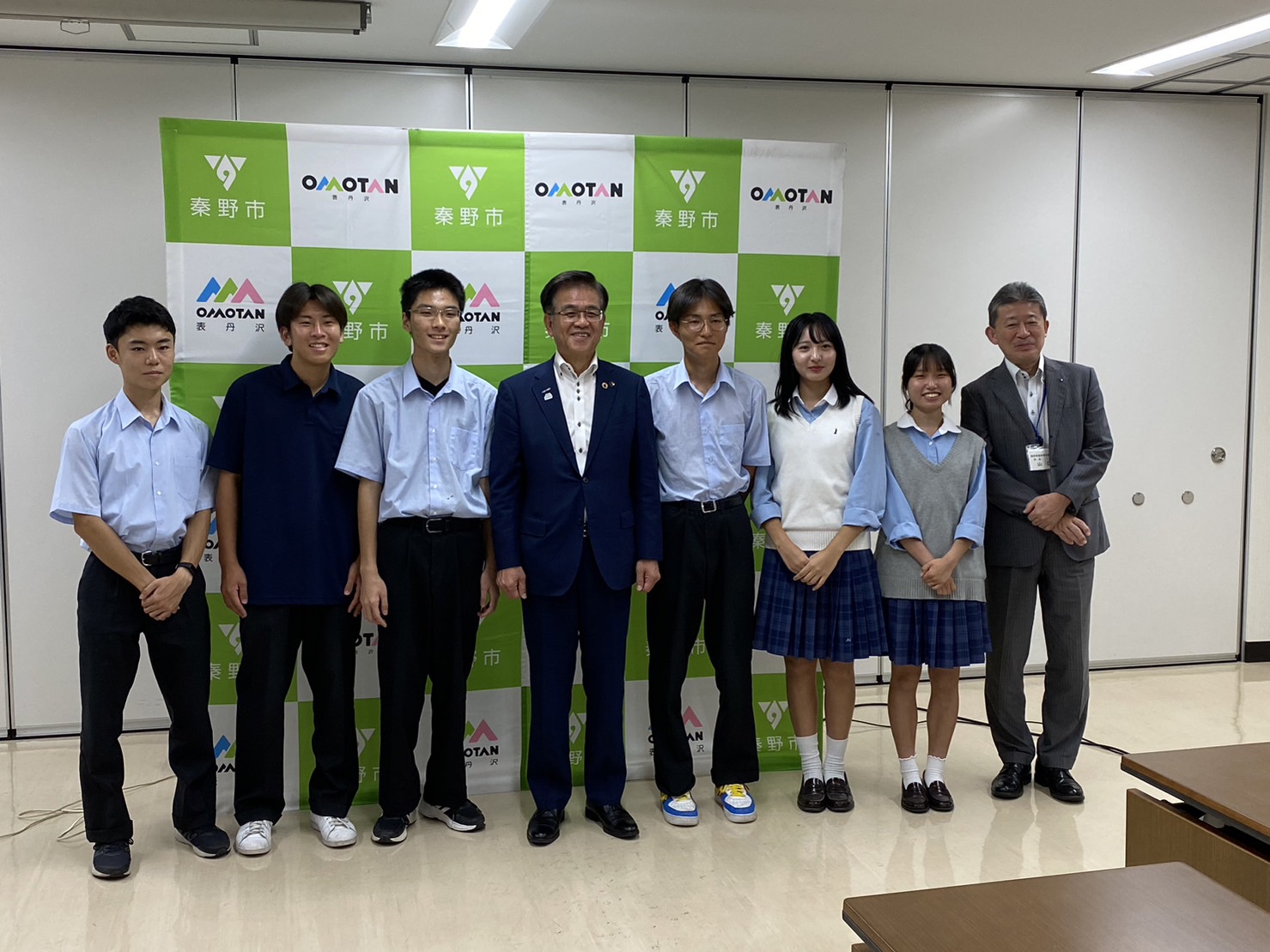 複数の学生と大人が、「秦野市」と「OMOTAN」のロゴがデザインされた緑と白のパネルの前で、記念撮影をしている集合写真