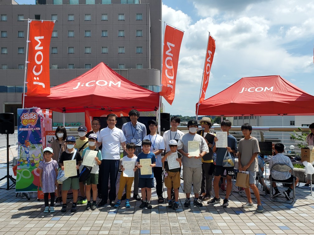 屋外イベント会場で、赤いJ:COMのテントと旗を背景に、子供から大人まで複数の人々が賞状を持ってカメラに向かって並んでいる集合写真