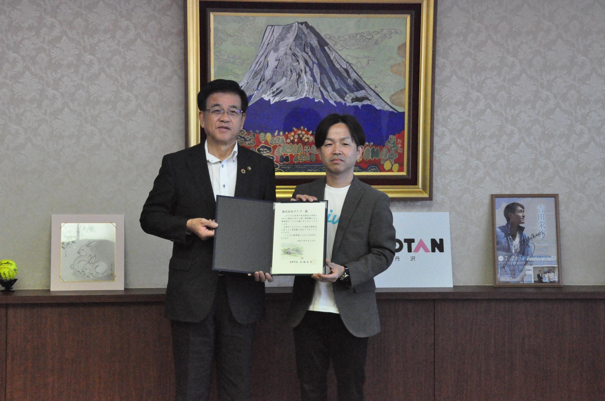 背景に富士山の絵画が飾られた部屋で、スーツ姿の市長とカジュアルなジャケット姿の男性の二人が、賞状のような書類を持って並んでいる様子の写真