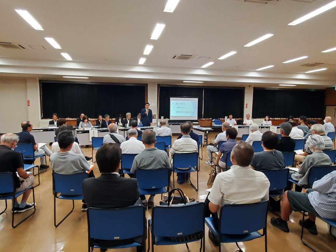 広い室内の奥にスクリーンがあり、その横の長机に関係者の方々が座り、中央のスーツ姿の市長がパイプ椅子に座っている参加者の人達に説明をしている写真