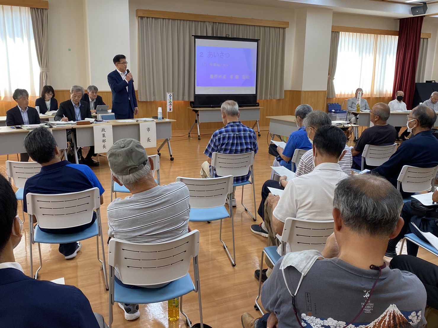 スクリーンの横の長机の席で、市長がマイクを持ちながら説明をし、パイプ椅子に座っている人たちが聞いている市政懇談会の写真