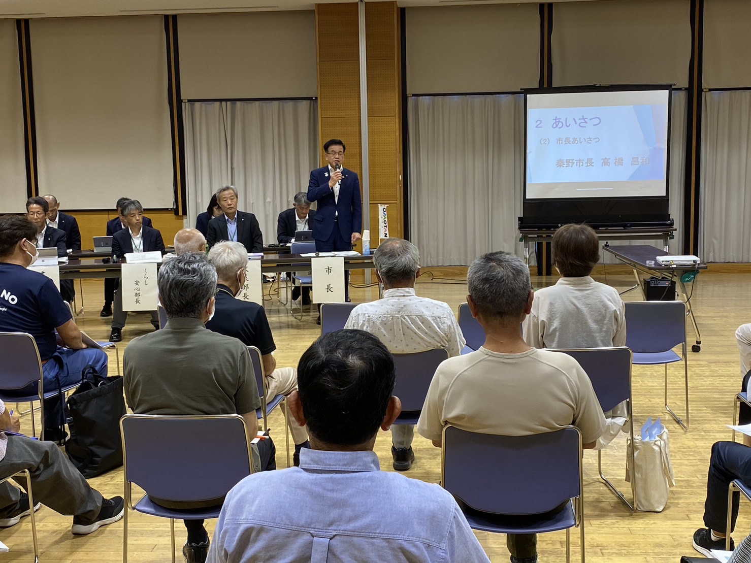 多くの人々が座って話を聞いている中、スクリーンに「あいさつ」と映し出された部屋で、前に立つスーツ姿の市長がマイクでスピーチをしている様子の写真