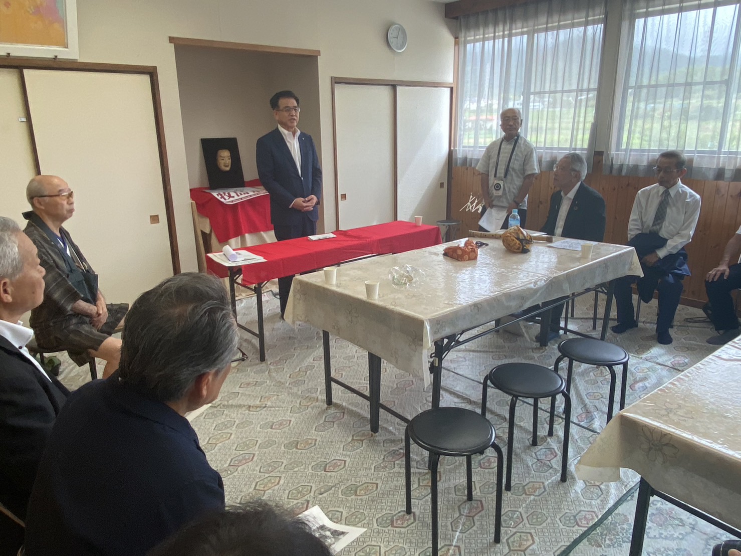 地域の集会室でスーツの市長が前に立ち、能面と赤い布が置かれた長机、その前の大きな机に紙コップや果物が並び、周囲に高齢の男性数人が座っている供養会での写真