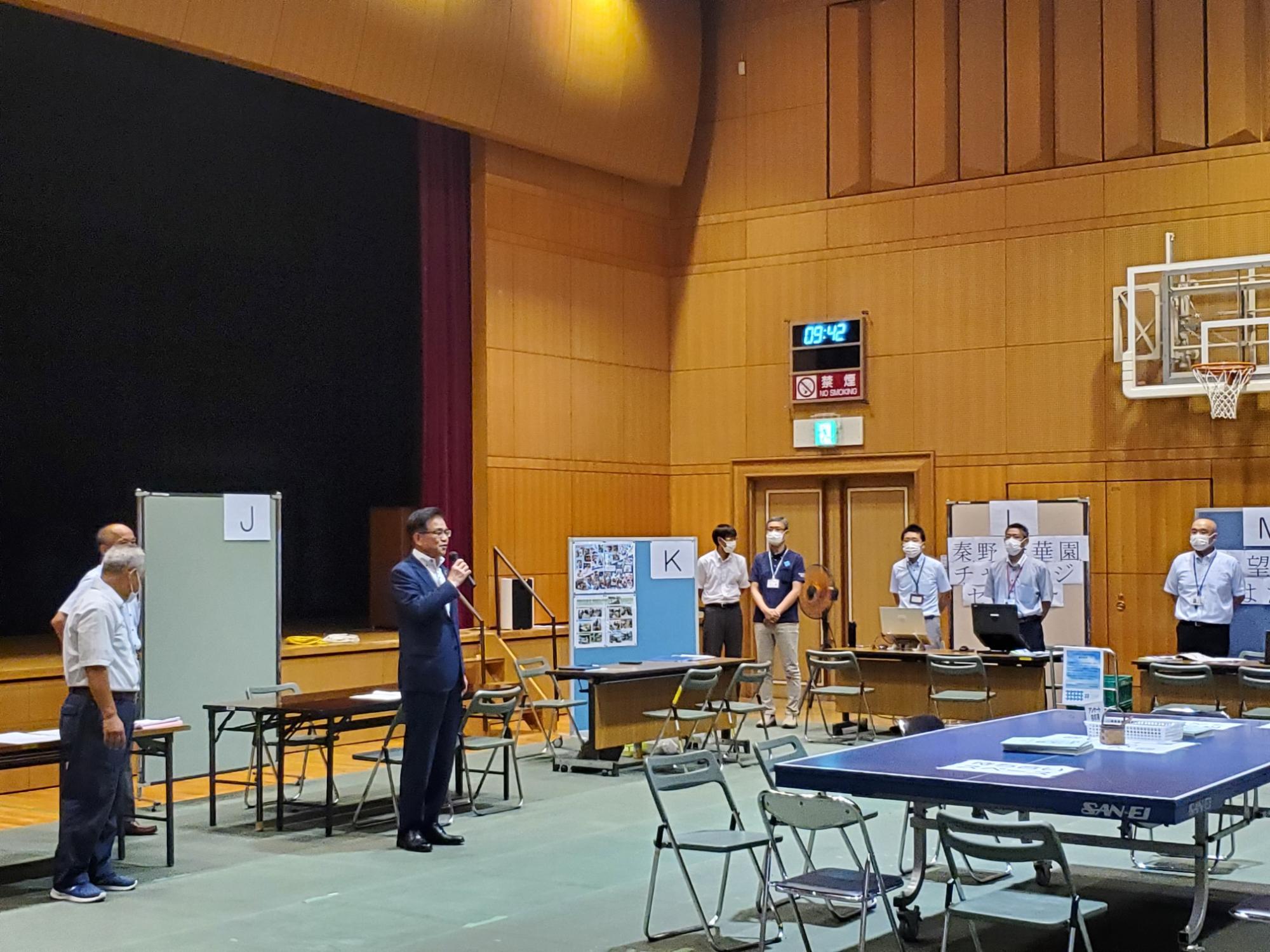 体育館のステージ前で、スーツ姿の市長がマイクでスピーチをしており、周りには多くの関係者と机と椅子などの設備が置かれている様子の写真