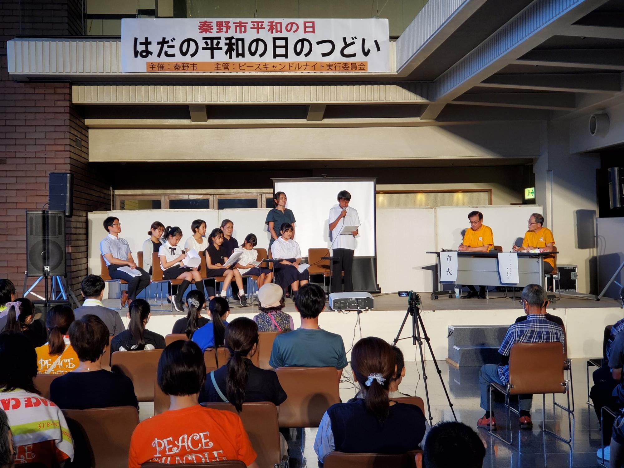 ステージ上で学生や大人たちが座っていて、モニターの前の男性がマイクで発表を行い、それを客席にいる多くの人々が聞いている様子の写真