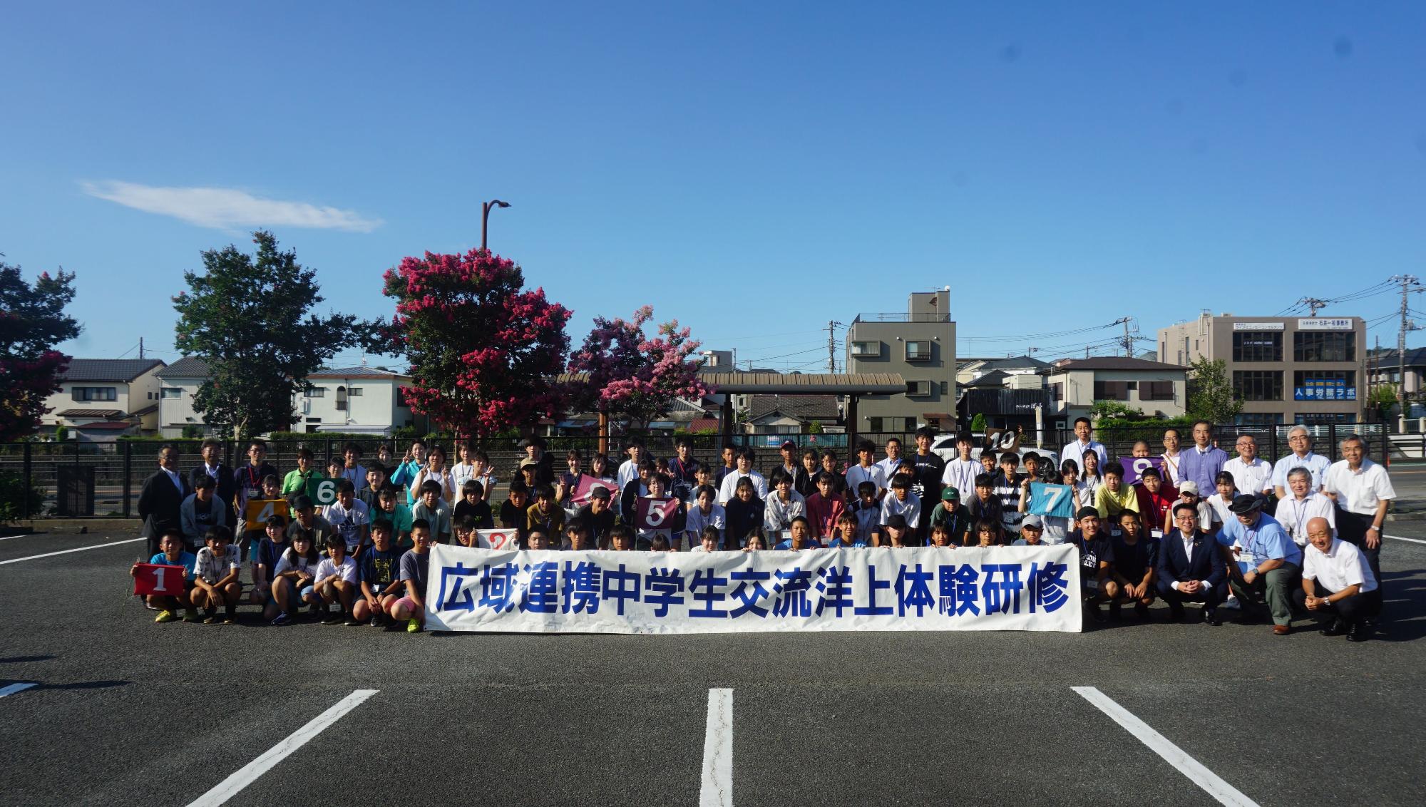 たくさんの子供たちと大人たちが、広々としたアスファルトの駐車場で、「広域連携中学生交流洋上体験研修」と書かれた横断幕を掲げて並んでいる集合写真