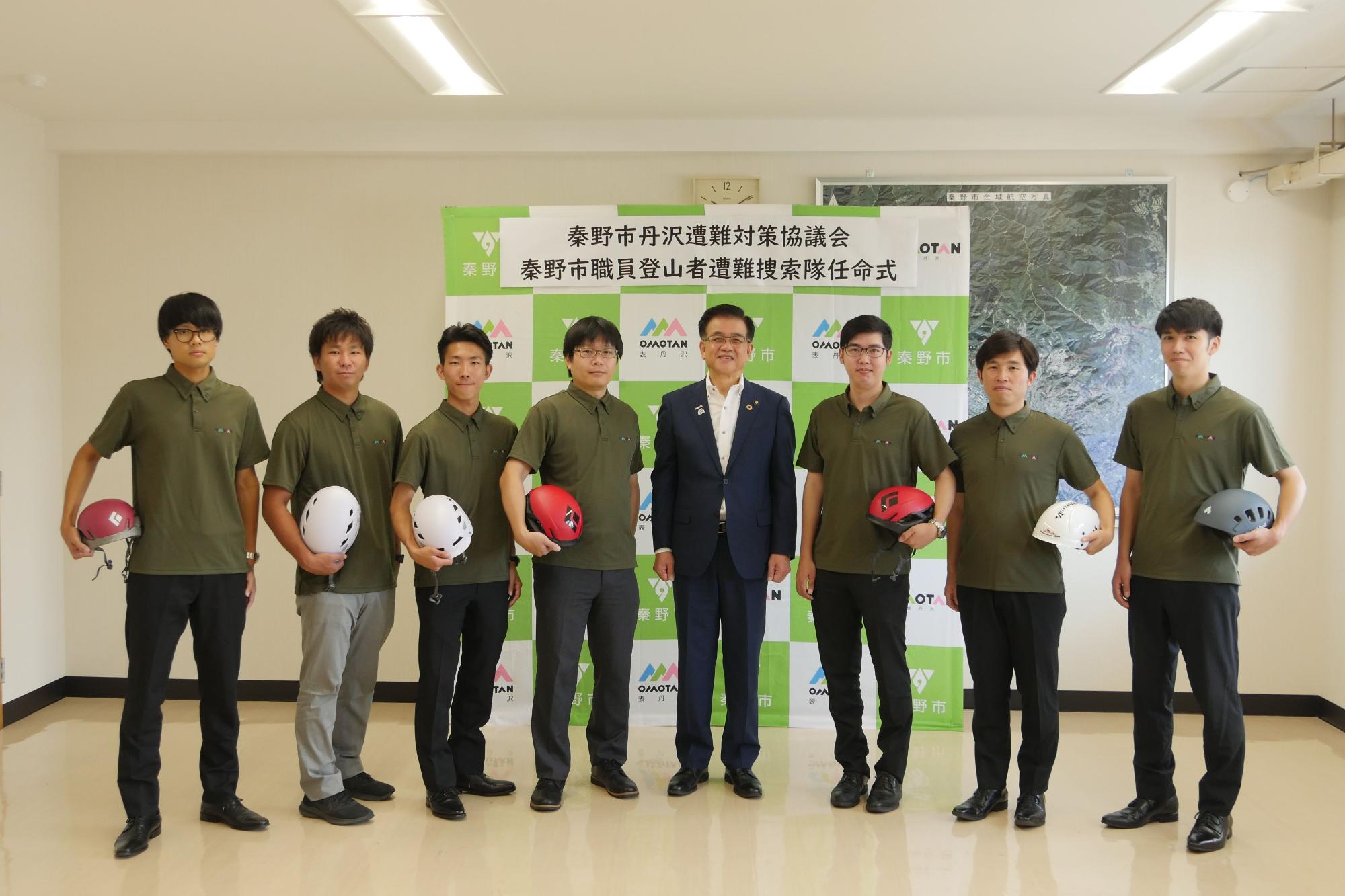 和やかな雰囲気の室内で、緑色のポロシャツを着用した7人の男性とスーツ姿の市長が、登山用ヘルメットを持って並んでいる集合写真