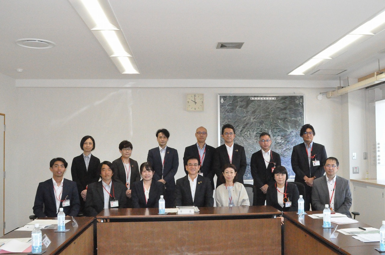 スーツ姿の男女14人が、U字型に配置された会議机を囲んで立ち、中央に座っている7人、立っている7人が共にカメラ目線で写っている集合写真