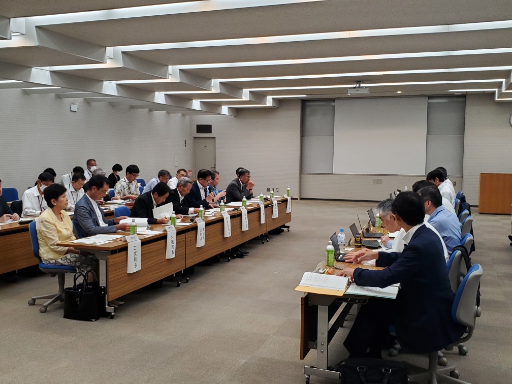 細長い会議室で、向かい合わせに机に座った複数の人物が真剣な表情で資料に目を通し、会議に参加している様子の写真